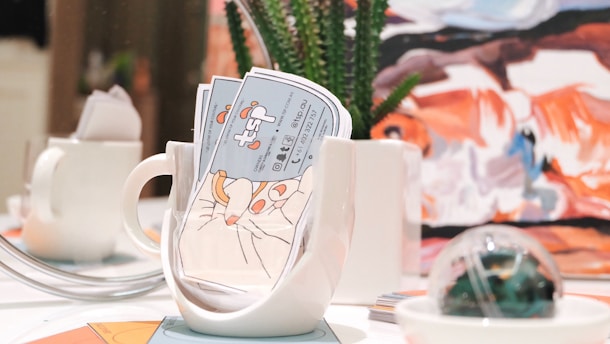 A brightly lit table setting features a white mug filled with colorful business cards, alongside a small white teapot. In the background, there is a piece of abstract art with bold strokes of orange, blue, and white. A succulent plant in a white pot and a decorative glass orb are also present on the table.