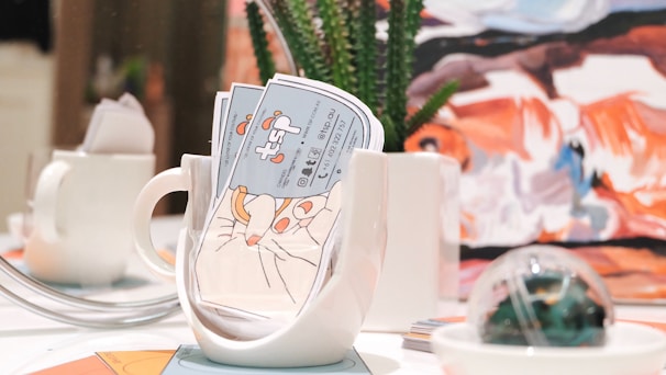 A brightly lit table setting features a white mug filled with colorful business cards, alongside a small white teapot. In the background, there is a piece of abstract art with bold strokes of orange, blue, and white. A succulent plant in a white pot and a decorative glass orb are also present on the table.