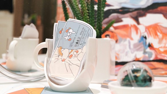 A brightly lit table setting features a white mug filled with colorful business cards, alongside a small white teapot. In the background, there is a piece of abstract art with bold strokes of orange, blue, and white. A succulent plant in a white pot and a decorative glass orb are also present on the table.