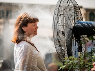 A woman stands in front of a large misting fan, enjoying a cooling spray of water droplets. She appears relaxed, dressed in a white blouse, with her eyes closed. The scene suggests a warm day, with sunlight casting shadows on the woman and the fan.