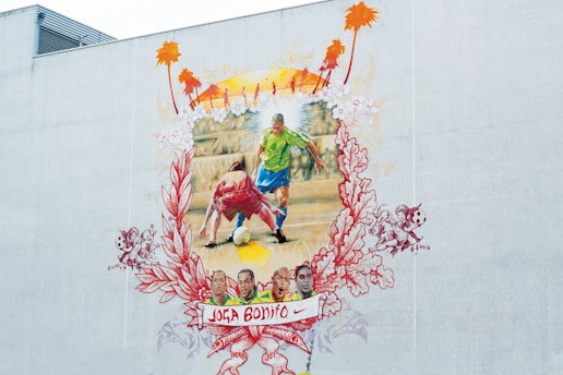 A colorful mural depicting a soccer scene where a player in a green and blue uniform moves past an opponent in a red and white uniform. The background includes a yellow and orange sunset with silhouettes of palm trees. Surrounding the central image, intricate floral and playful designs frame the scene. At the bottom, a banner with the words 'Joga Bonito' and a Nike logo is visible, along with several smaller images of faces and soccer-related motifs.