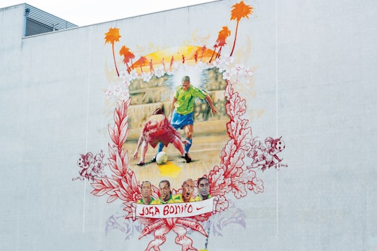 A colorful mural depicting a soccer scene where a player in a green and blue uniform moves past an opponent in a red and white uniform. The background includes a yellow and orange sunset with silhouettes of palm trees. Surrounding the central image, intricate floral and playful designs frame the scene. At the bottom, a banner with the words 'Joga Bonito' and a Nike logo is visible, along with several smaller images of faces and soccer-related motifs.