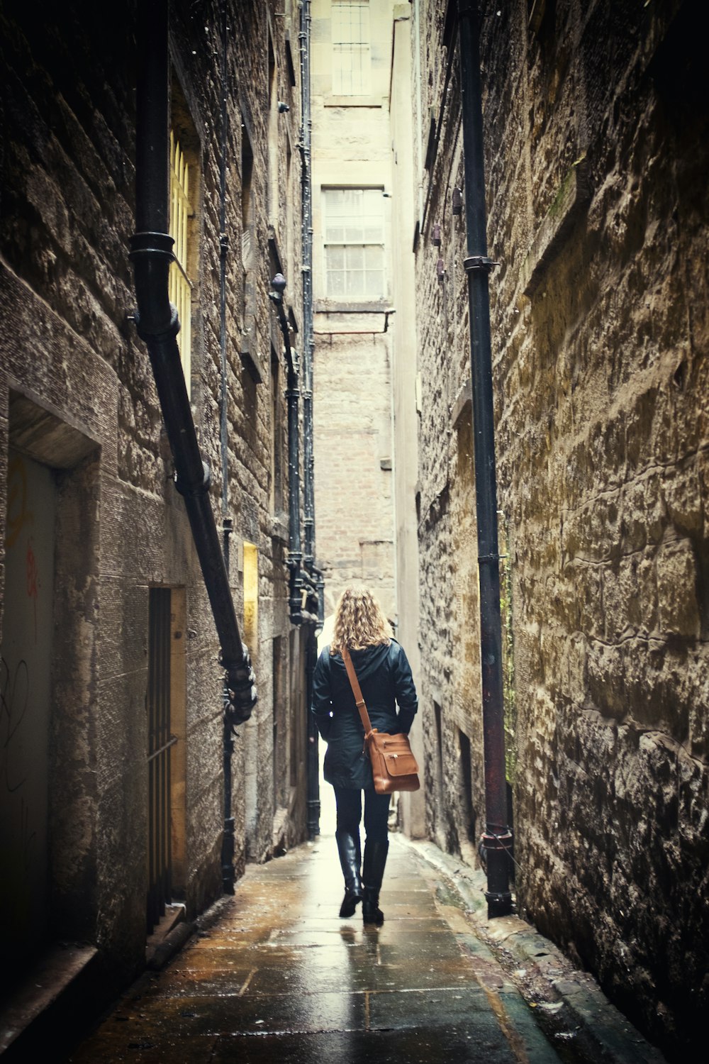 woman carrying brown leather shoulder bag walking on narrow concrete pathway between concrete buildings during daytime