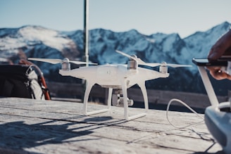 A white drone is positioned on a wooden surface outdoors with a backdrop of snow-covered mountains. A person is holding a remote control device to the right, possibly operating the drone.