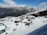A snowy mountain landscape with several small buildings and a group of people trekking or skiing through the snow. The foreground features sloped, snow-covered terrain leading down to the buildings, which have brightly colored roofs. In the background, majestic mountains with snow-capped peaks rise against a clear blue sky with scattered clouds.