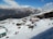 A snowy mountain landscape with several small buildings and a group of people trekking or skiing through the snow. The foreground features sloped, snow-covered terrain leading down to the buildings, which have brightly colored roofs. In the background, majestic mountains with snow-capped peaks rise against a clear blue sky with scattered clouds.