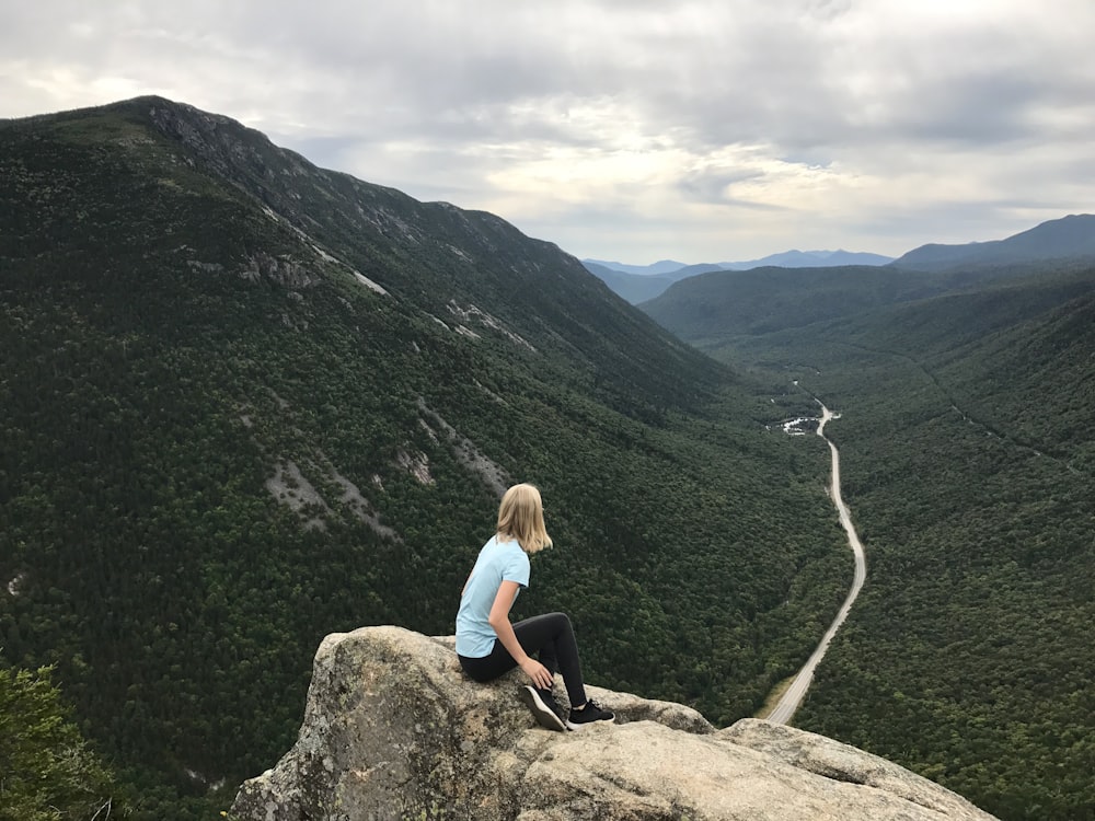 woman in blue t-shirt sitting on brown rock facing green mountains