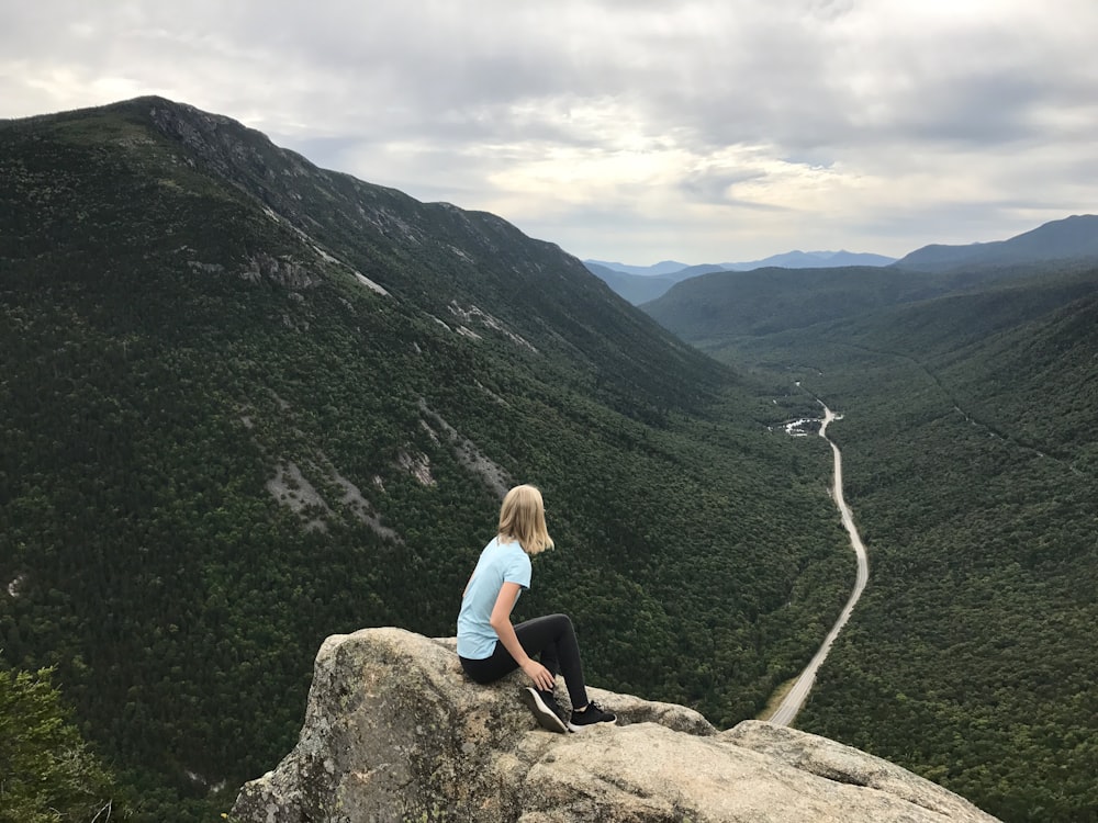 Mujer con camiseta azul sentada en roca marrón frente a montañas verdes