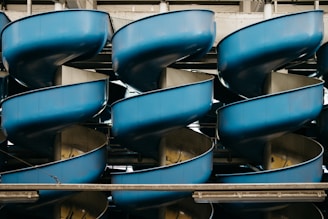 Several intertwined, circular blue slides stacked vertically within a metallic structure give an industrial and geometric appearance. The slides create a repeating pattern and seem to be part of an amusement or water park.