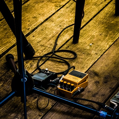 A wooden stage floor with various audio equipment, including a yellow guitar effects pedal with cables attached. Metal stands or parts possibly supporting instruments are visible, along with a microphone stand.
