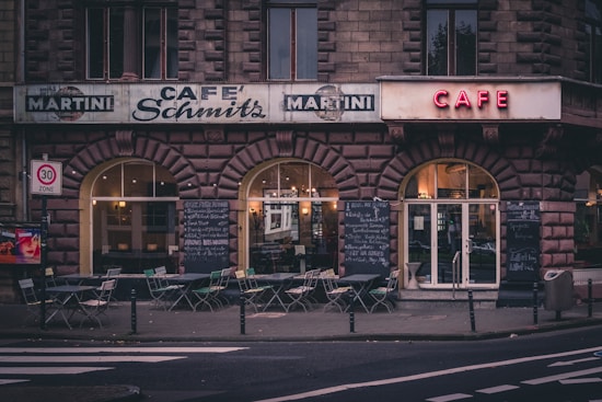 A vintage-style caf&eacute; with a brick facade and arched windows, displaying several signs including 'Caf&eacute; Schmitz' and 'Martini'. The outdoor area is equipped with empty tables and chairs, and there are large chalkboards displaying menus on either side of the entrance. The atmosphere suggests early evening or late afternoon, with dim lighting inside the caf&eacute;.