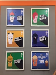 A display of six framed posters on a grey wall featuring various graphic designs and text related to milkshakes. Each poster has a colorful background and an illustration resembling a character with words like 'I just love the science of shakes,' 'sexy shakes,' and 'thickshake? oh yeah.' The posters have a humorous and playful design with vibrant characters.