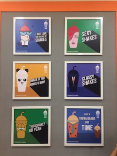 A display of six framed posters on a grey wall featuring various graphic designs and text related to milkshakes. Each poster has a colorful background and an illustration resembling a character with words like 'I just love the science of shakes,' 'sexy shakes,' and 'thickshake? oh yeah.' The posters have a humorous and playful design with vibrant characters.