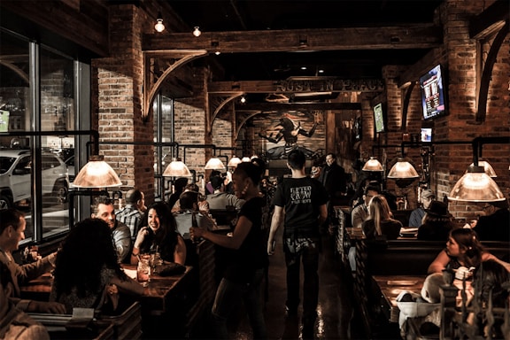 A lively bar or restaurant setting with groups of people seated in wooden booths, engaging in conversations and enjoying their time. The interior features exposed brick walls, industrial-style lighting, and multiple television screens mounted on the walls. The atmosphere appears warm and cozy, with dim lighting casting a welcoming glow.