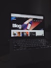 A laptop displays a web page with a bold header text reading 'Blog' against a colorful abstract background. Several thumbnail images are displayed below the header, featuring various content such as graphics and text. The screen is illuminated, creating a contrast against the dark keyboard and surrounding environment.