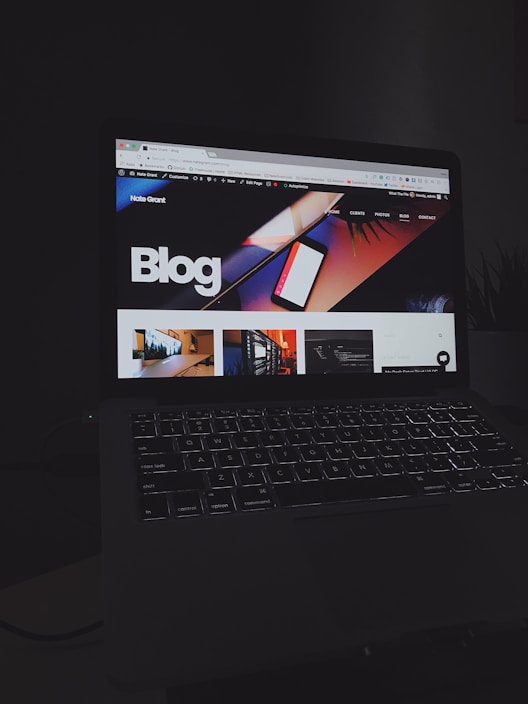 A laptop displays a web page with a bold header text reading 'Blog' against a colorful abstract background. Several thumbnail images are displayed below the header, featuring various content such as graphics and text. The screen is illuminated, creating a contrast against the dark keyboard and surrounding environment.