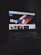 A laptop displays a web page with a bold header text reading 'Blog' against a colorful abstract background. Several thumbnail images are displayed below the header, featuring various content such as graphics and text. The screen is illuminated, creating a contrast against the dark keyboard and surrounding environment.