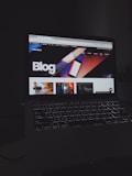 A laptop displays a web page with a bold header text reading 'Blog' against a colorful abstract background. Several thumbnail images are displayed below the header, featuring various content such as graphics and text. The screen is illuminated, creating a contrast against the dark keyboard and surrounding environment.