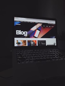 A laptop displays a web page with a bold header text reading 'Blog' against a colorful abstract background. Several thumbnail images are displayed below the header, featuring various content such as graphics and text. The screen is illuminated, creating a contrast against the dark keyboard and surrounding environment.