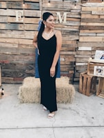 A woman in a black evening gown stands in front of a wooden pallet backdrop. She is adorned with a necklace and earrings, and has her hair styled fashionably. Hay bales and crates are positioned around, contributing to a rustic theme. String lights hang above, adding a warm ambiance.