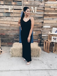 A woman in a black evening gown stands in front of a wooden pallet backdrop. She is adorned with a necklace and earrings, and has her hair styled fashionably. Hay bales and crates are positioned around, contributing to a rustic theme. String lights hang above, adding a warm ambiance.