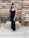 A woman in a black evening gown stands in front of a wooden pallet backdrop. She is adorned with a necklace and earrings, and has her hair styled fashionably. Hay bales and crates are positioned around, contributing to a rustic theme. String lights hang above, adding a warm ambiance.