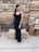 A woman in a black evening gown stands in front of a wooden pallet backdrop. She is adorned with a necklace and earrings, and has her hair styled fashionably. Hay bales and crates are positioned around, contributing to a rustic theme. String lights hang above, adding a warm ambiance.