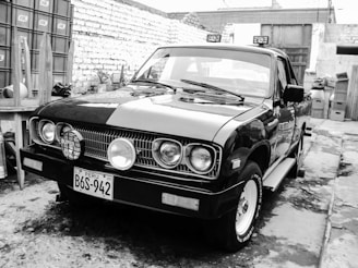 A classic pickup truck with a distinctive front grille and circular headlights parked in an outdoor setting. The vehicle has a Peruvian license plate and is situated against a backdrop of brick walls and assorted household items.