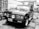 A classic pickup truck with a distinctive front grille and circular headlights parked in an outdoor setting. The vehicle has a Peruvian license plate and is situated against a backdrop of brick walls and assorted household items.
