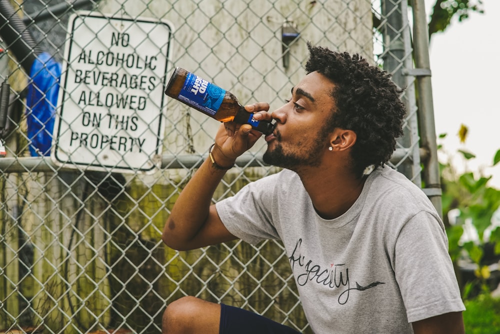 man drinking beverage in amber glass bottle beside gray wire link fence at daytime