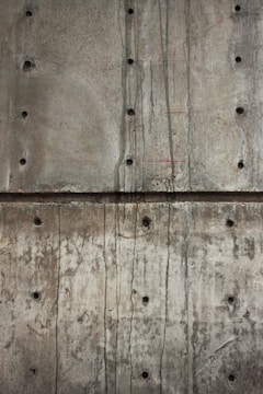 A textured concrete wall with visible holes possibly left from formwork and dark streaks of water or stains running vertically. The surface shows various shades of gray and brown, indicating wear and aging.