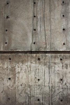A textured concrete wall with visible holes possibly left from formwork and dark streaks of water or stains running vertically. The surface shows various shades of gray and brown, indicating wear and aging.