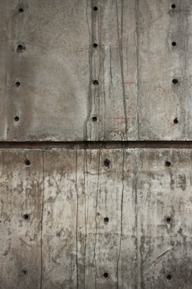 A textured concrete wall with visible holes possibly left from formwork and dark streaks of water or stains running vertically. The surface shows various shades of gray and brown, indicating wear and aging.