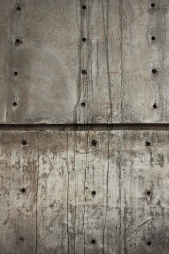 A textured concrete wall with visible holes possibly left from formwork and dark streaks of water or stains running vertically. The surface shows various shades of gray and brown, indicating wear and aging.