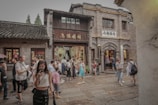 A bustling street in what appears to be a traditional area, with people walking and taking photos. The buildings have an old architectural style, with brick walls and wooden details, likely from a historical or cultural site. The presence of signs with Asian characters suggests a location in East Asia. The scene is lively with tourists and visitors exploring the area.