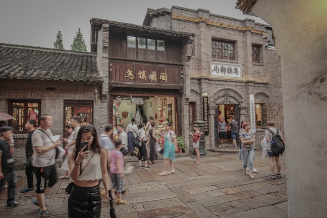 A bustling street in what appears to be a traditional area, with people walking and taking photos. The buildings have an old architectural style, with brick walls and wooden details, likely from a historical or cultural site. The presence of signs with Asian characters suggests a location in East Asia. The scene is lively with tourists and visitors exploring the area.