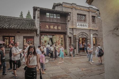 A bustling street in what appears to be a traditional area, with people walking and taking photos. The buildings have an old architectural style, with brick walls and wooden details, likely from a historical or cultural site. The presence of signs with Asian characters suggests a location in East Asia. The scene is lively with tourists and visitors exploring the area.