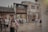 A bustling street in what appears to be a traditional area, with people walking and taking photos. The buildings have an old architectural style, with brick walls and wooden details, likely from a historical or cultural site. The presence of signs with Asian characters suggests a location in East Asia. The scene is lively with tourists and visitors exploring the area.