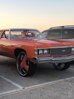 A classic orange car with oversized wheels is parked in a lot next to a gray SUV. The car features a chrome grille and vintage design elements. The background shows a clear sky at sunset, adding a warm glow to the scene.
