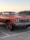 A classic orange car with oversized wheels is parked in a lot next to a gray SUV. The car features a chrome grille and vintage design elements. The background shows a clear sky at sunset, adding a warm glow to the scene.