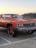 A classic orange car with oversized wheels is parked in a lot next to a gray SUV. The car features a chrome grille and vintage design elements. The background shows a clear sky at sunset, adding a warm glow to the scene.