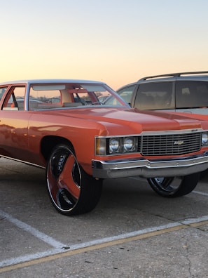 A classic orange car with oversized wheels is parked in a lot next to a gray SUV. The car features a chrome grille and vintage design elements. The background shows a clear sky at sunset, adding a warm glow to the scene.