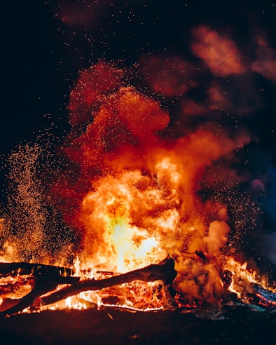 Bright flames and smoke fill the scene, with sparks flying against a dark background. Large logs and burning debris are engulfed in the fire, creating an intense and dynamic visual effect.