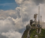 A rocky mountaintop with lush green vegetation hosts various weather monitoring or communication equipment. Large, dome-shaped structures and antennas are visible, surrounded by dramatic clouds and a partly cloudy blue sky.
