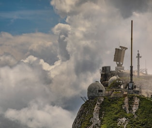 A rocky mountaintop with lush green vegetation hosts various weather monitoring or communication equipment. Large, dome-shaped structures and antennas are visible, surrounded by dramatic clouds and a partly cloudy blue sky.