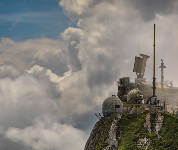A rocky mountaintop with lush green vegetation hosts various weather monitoring or communication equipment. Large, dome-shaped structures and antennas are visible, surrounded by dramatic clouds and a partly cloudy blue sky.
