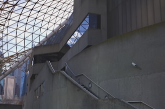 A modern architectural structure with geometric designs, featuring a triangular glass roof supported by metal beams. The concrete walls rise up with steps and a railing leading upwards. A section of the city skyline is visible in the background, offering a glimpse of high-rise buildings. A security camera is mounted on one of the walls.