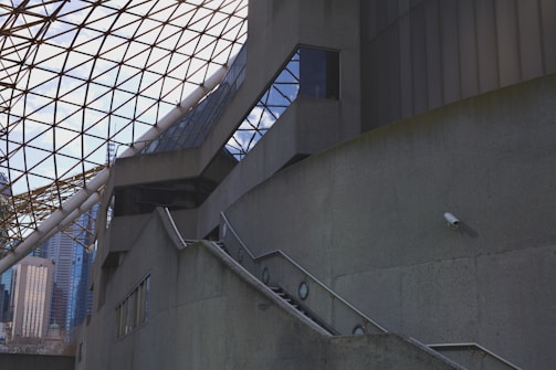 A modern architectural structure with geometric designs, featuring a triangular glass roof supported by metal beams. The concrete walls rise up with steps and a railing leading upwards. A section of the city skyline is visible in the background, offering a glimpse of high-rise buildings. A security camera is mounted on one of the walls.