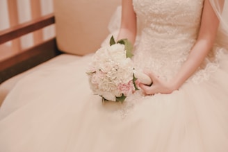 A bride is seated, holding a bouquet of white and pale pink flowers. The intricate lace and beading on her wedding dress are visible, and the background has a soft, blurry effect.