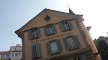 A European-style building with beige walls and multiple windows featuring blue shutters. The text 'Restaurant Beaulieu' is prominently displayed. The building has a triangular roof with an architectural detail near the top, possibly a small circular window or decoration. This establishment is situated in an area with additional modern buildings visible in the background.